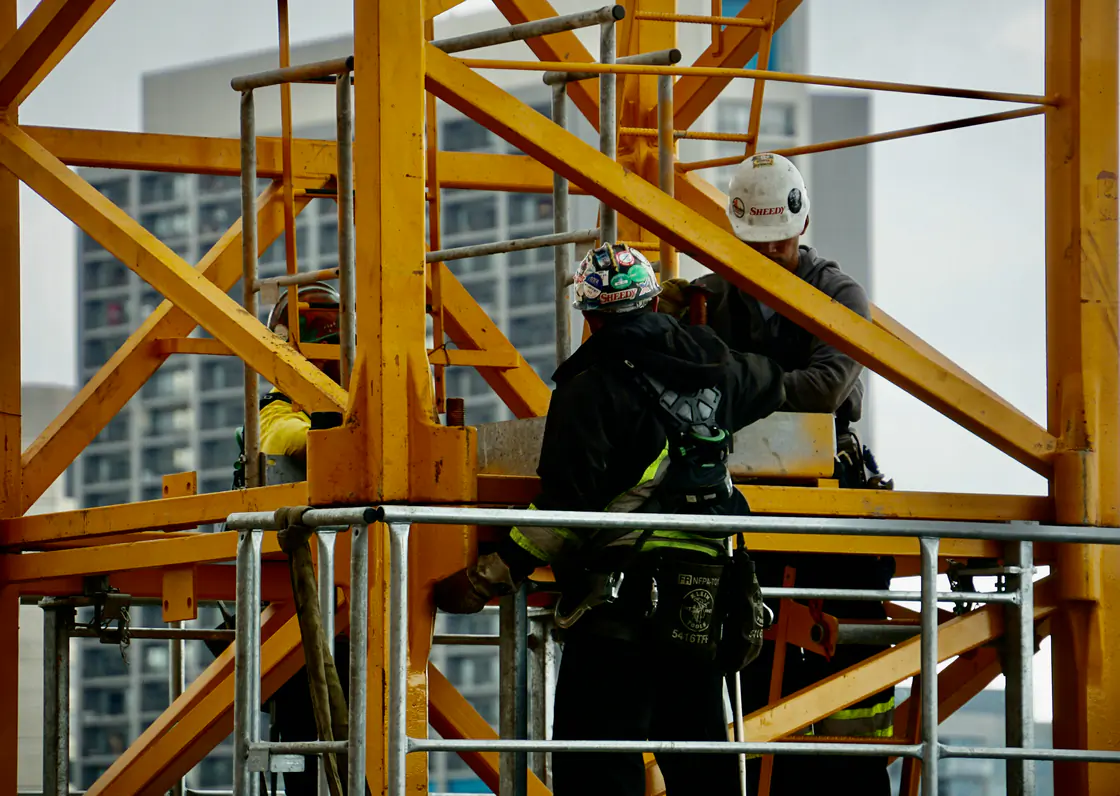 Two workers in hard hats and full-body harnesses working inside the guardrails of a yellow tower-crane section, high over the city.